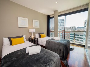 A hotel room with two beds, a large window with a city view, and artwork on the wall.