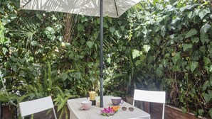Um pátio externo com mesa branca, duas cadeiras brancas, guarda-sol de mesa e diversas plantas ao fundo.
