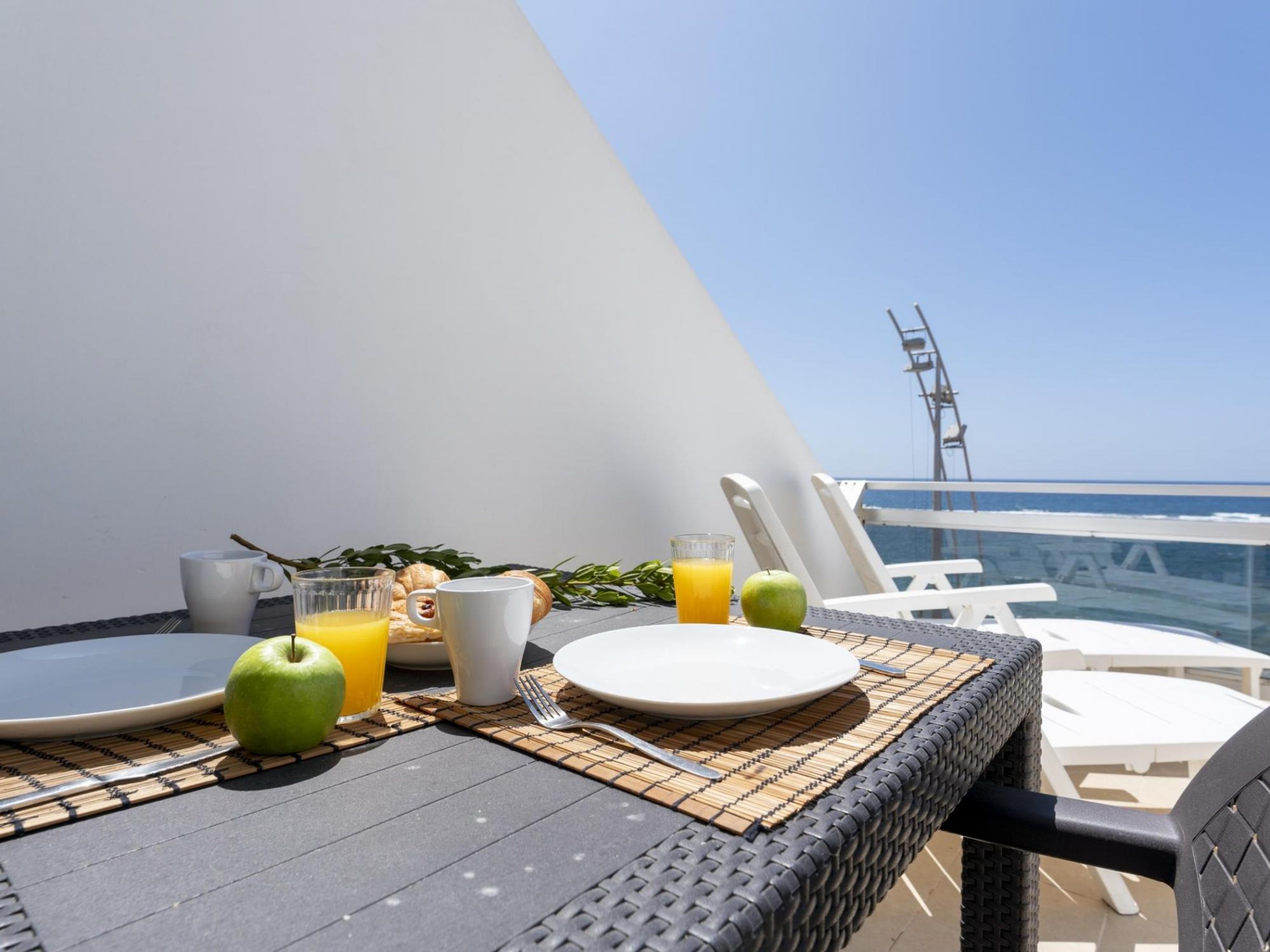 Mesa posta com pratos, um copo de suco de laranja, uma caneca branca e uma maçã, com vista para o mar e uma roda gigante ao fundo.
