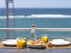 A breakfast setup with croissants, juice, and a bottle on a table with a view of the ocean and a bridge. - Apartamento Mars n Las Canteras Vista mar 308 (Las Palmas de Gran Canaria)