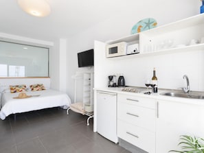 Ein modernes, kompaktes Schlafzimmer mit Bett, Nachttisch und einem großen Fenster. An das Schlafzimmer angrenzend befindet sich eine Küchenzeile mit Mikrowelle, Toaster und Spüle.