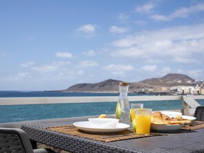 Ein Tisch mit Frühstücksangeboten und einer Wasserflasche, mit Blick auf die Küste und Berge im Hintergrund.