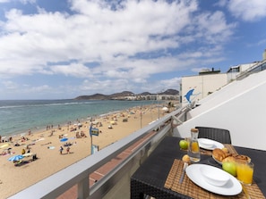 A balcony with a table set for breakfast overlooking a crowded beach with umbrellas and people.