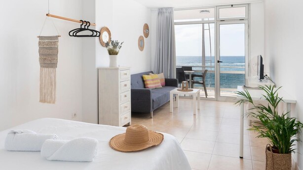 A modern hotel room with a balcony, a bed with white linens, a small table, and a view of the sea.
