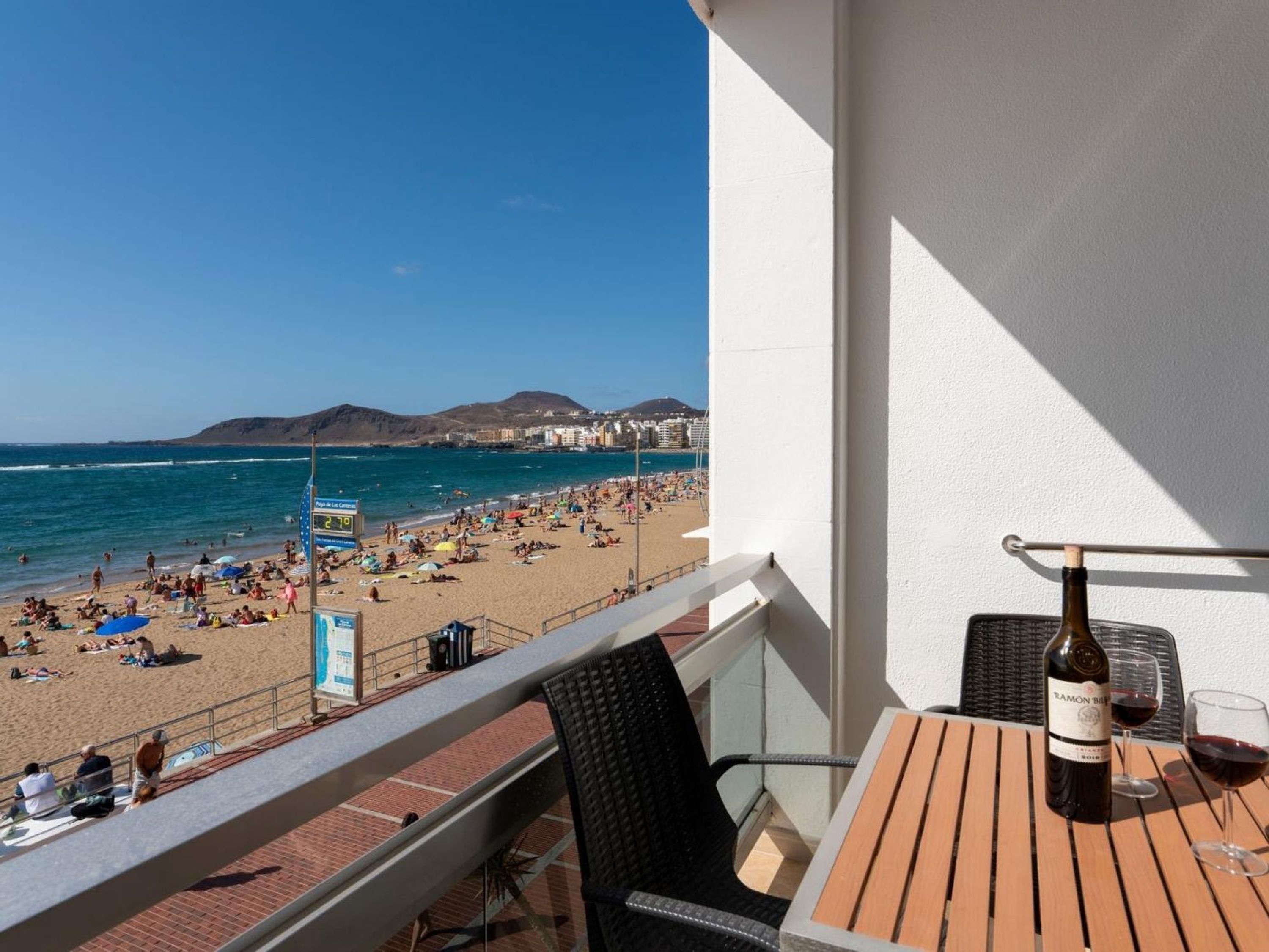 Varanda com mesa e cadeiras, com vista para uma praia lotada.