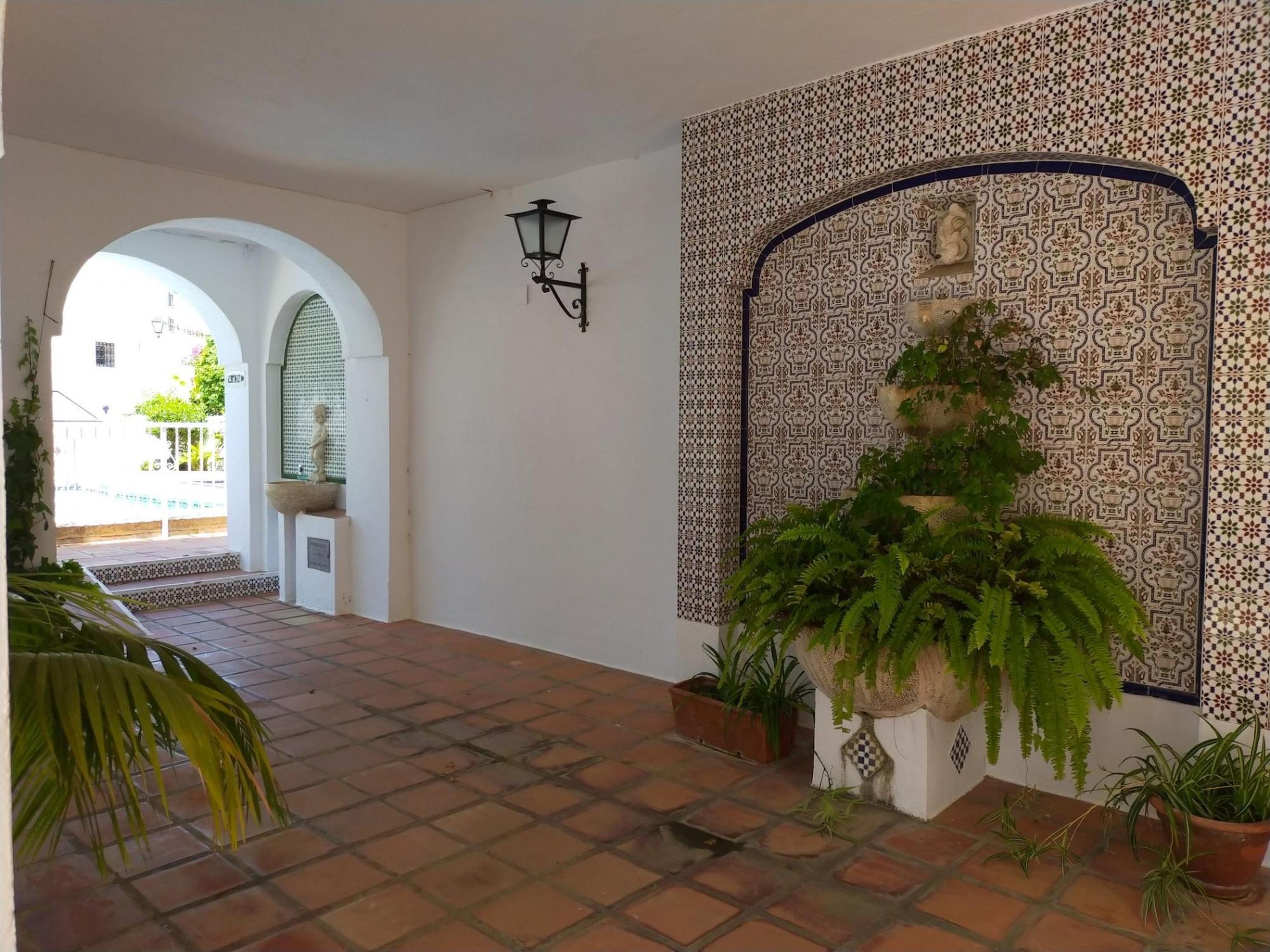 Corredor com piso de azulejos, paredes brancas e detalhes em mosaico na lateral direita. Há plantas em vasos e um aplique na parede.