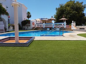 Una piscina con solárium de baldosas, una palmera y un edificio blanco con balcón.