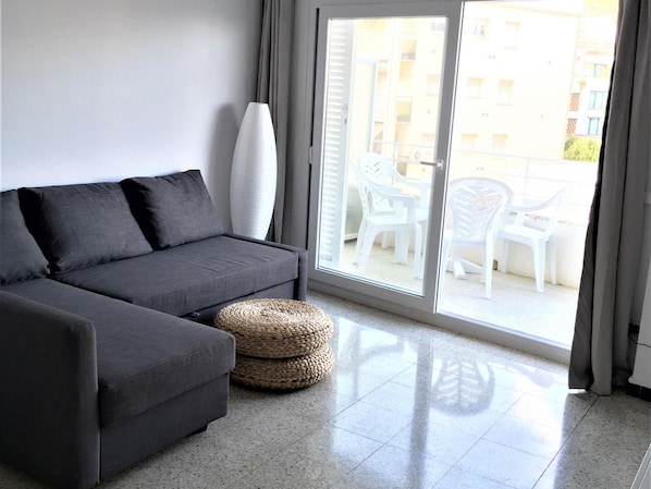 A modern living room with a grey sofa, a woven ottoman, and a white surfboard leaning against the wall. - Apartamento Costa Brava CON Vistas AL MAR (Torroella de Montgrí)
