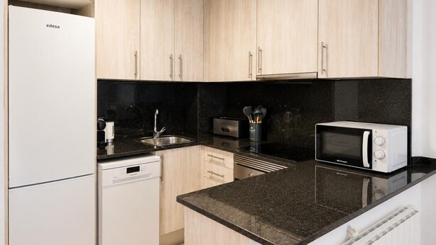 A modern kitchen with light wood cabinets, a black countertop, a dishwasher, a microwave, and a stove. - Apartament Acabat de Reformar a 1 min de la Platja (Palafrugell)
