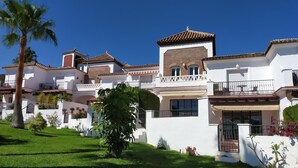 A multi-story residential building with white walls and terracotta roofs, featuring balconies and a well-maintained garden with palm trees and shrubs. - Casa Cipreses 4 Nerja (Nerja)