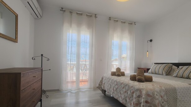 A bedroom with a bed, a wooden dresser, a mirror, and a window with curtains. - Casa Cipreses 4 Nerja (Nerja)