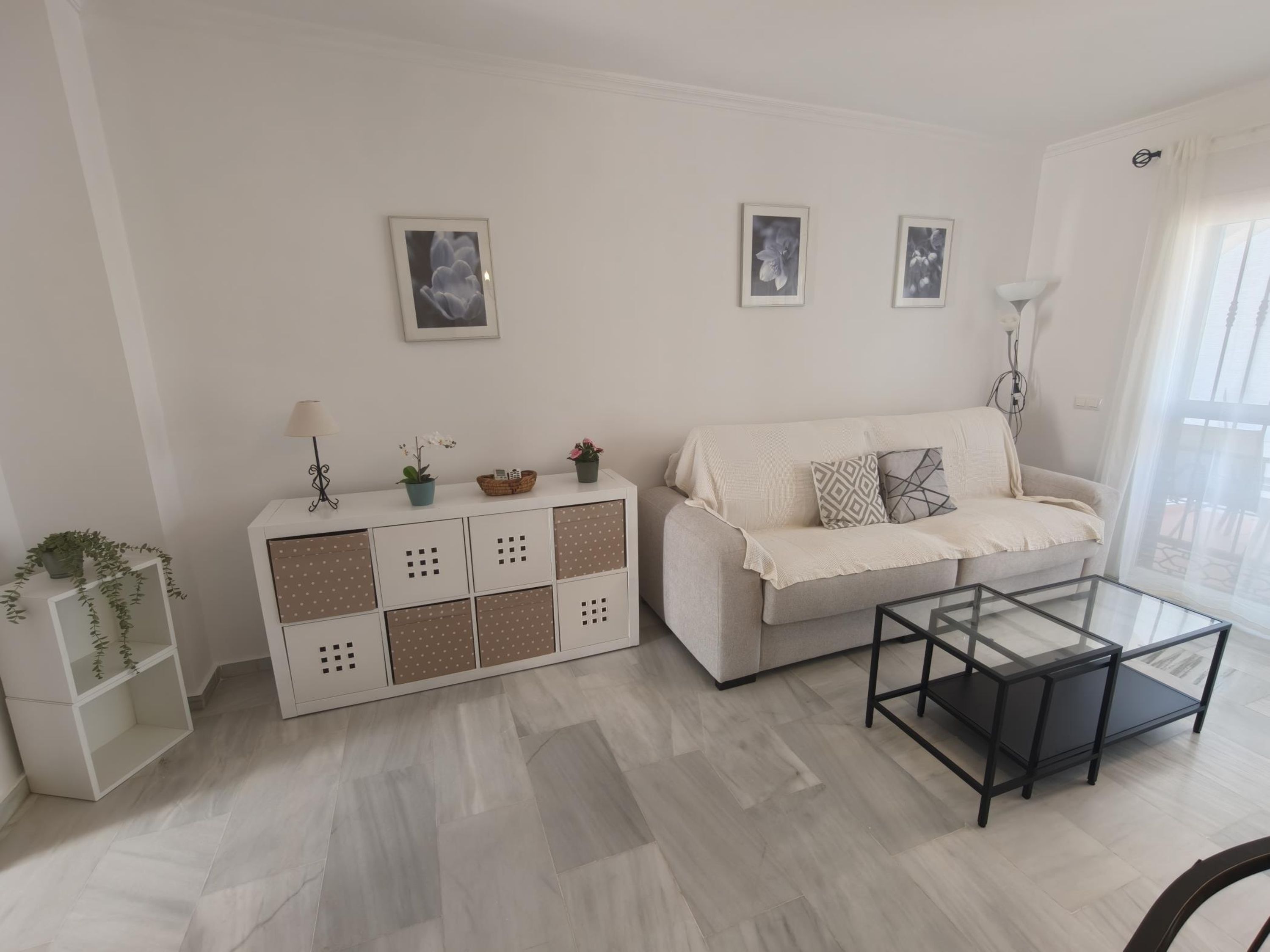 Ein modernes Wohnzimmer mit einem weißen Sofa, einem Glastisch und einer weißen Kommode mit dekorativen Verzierungen. An der Wand hängen drei Bilder und ein Stehlampen steht dort.