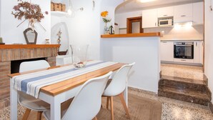 Un comedor con una mesa de madera y sillas blancas, chimenea de ladrillo y cocina con armarios blancos y horno empotrado.