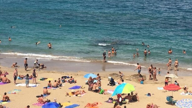 Una playa concurrida con gente nadando y tomando el sol, y sombrillas que ofrecen sombra.