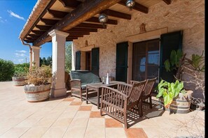 Un porche cubierto con una mesa y sillas de madera, paredes de piedra y plantas en macetas.