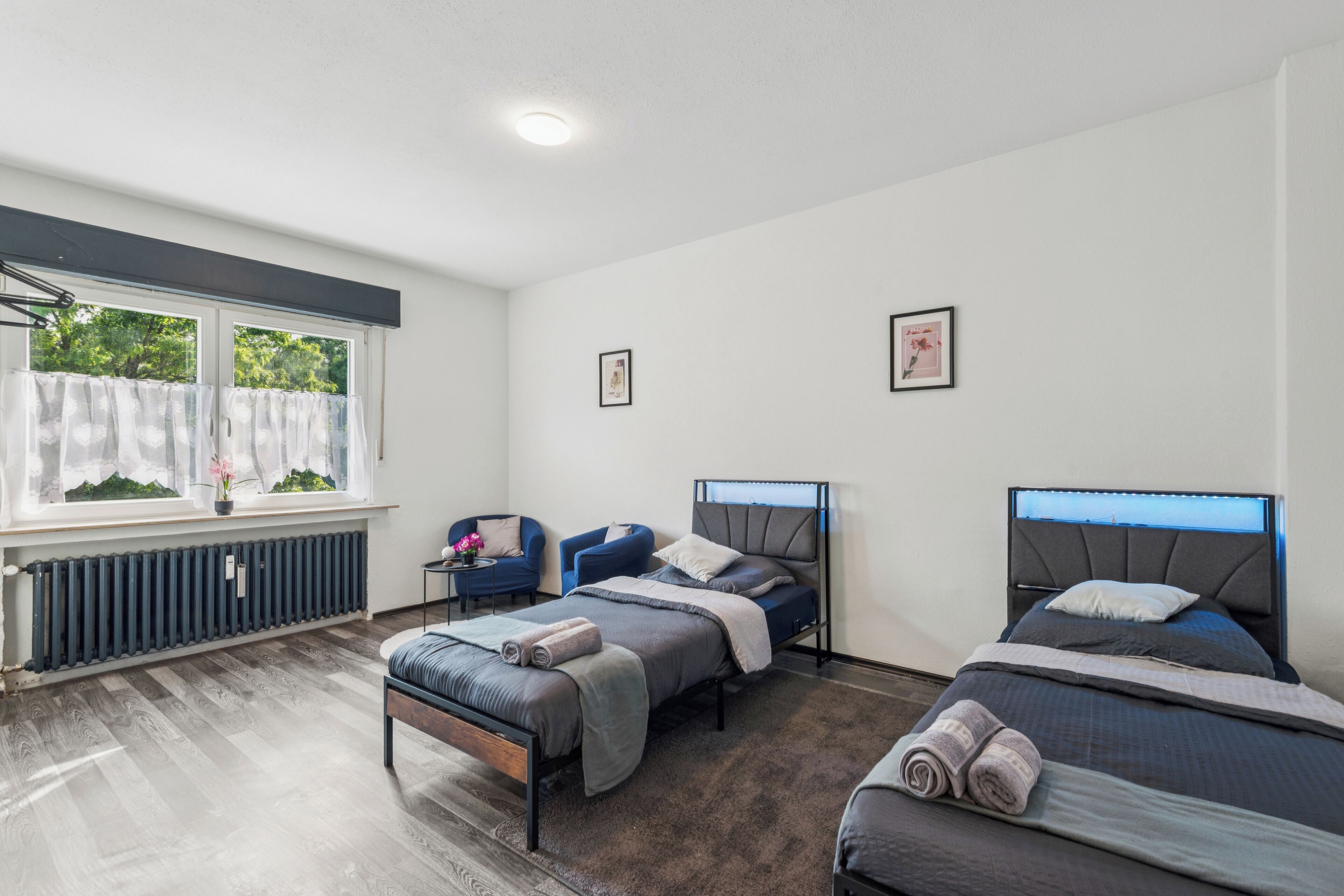 A bedroom with two beds, a window with curtains, a radiator, and a small table with a vase.