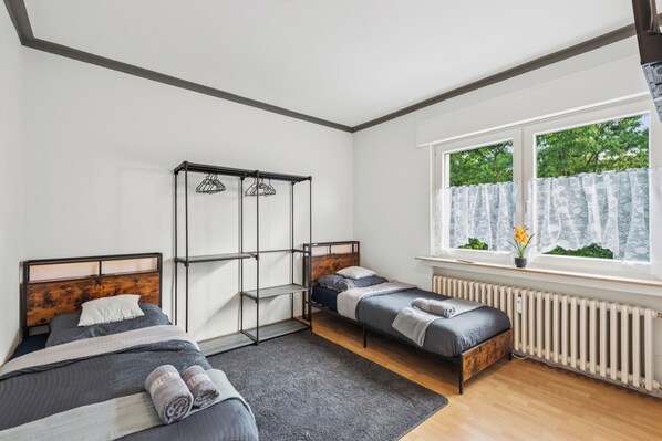 Ein Schlafzimmer mit zwei Betten, einem Metallschrank, einem Heizkörper und einem Fenster mit Blick auf Bäume.