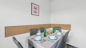 Comedor con una mesa dispuesta para cuatro, incluyendo platos, cubiertos y vasos. Hay dos botellas verdes y un frutero sobre la mesa. La habitación tiene una pared blanca con un cuadro enmarcado sobre la mesa.