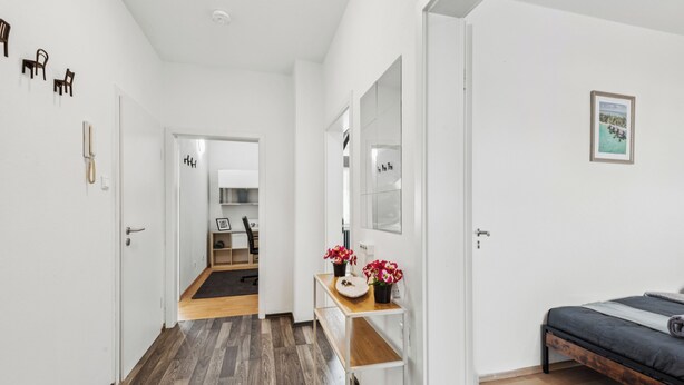 Un dormitorio moderno con suelo de madera, una cama, un escritorio con espejo y una pequeña mesa con flores.