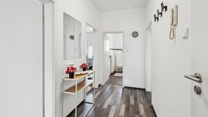 Un pasillo con una puerta blanca, un espejo, una estantería con flores, un teléfono y un reloj en la pared.