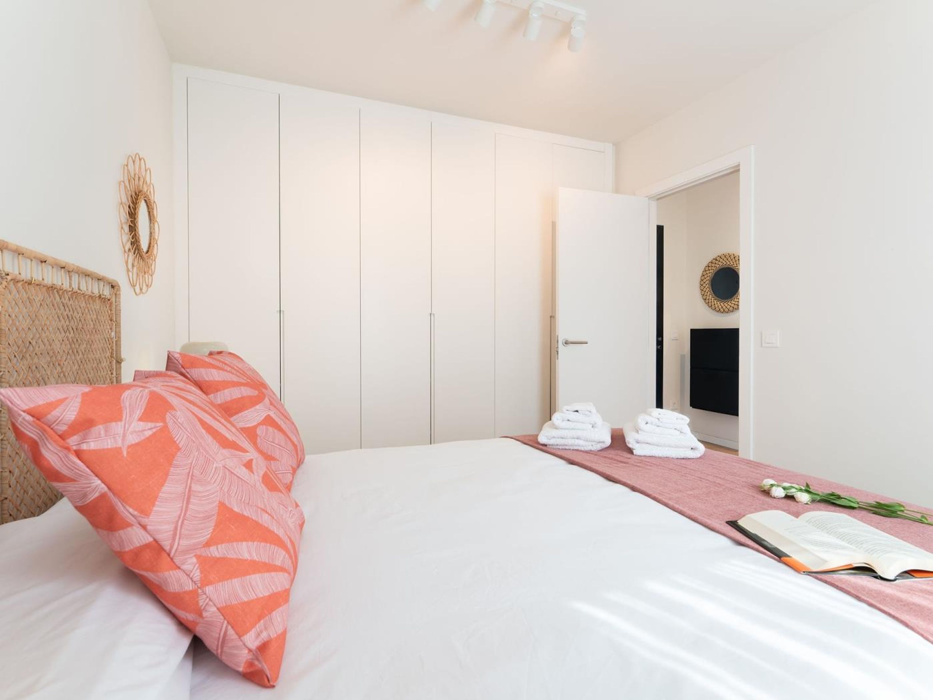 A neatly made bed with a white and orange patterned pillow, a pink bed runner, and a book on the side. There are two white towels stacked on the bed runner.