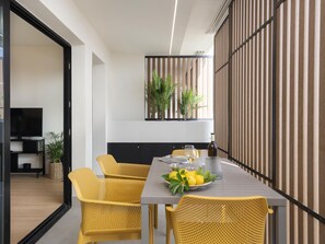 Een moderne eetruimte met een lange tafel, gele geweven stoelen en een decoratieve plant aan de muur.