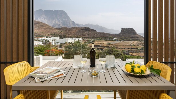 Une table avec une bouteille de vin, des verres, un bol de citrons et des magazines, le tout placé sur un balcon donnant sur un paysage montagneux.