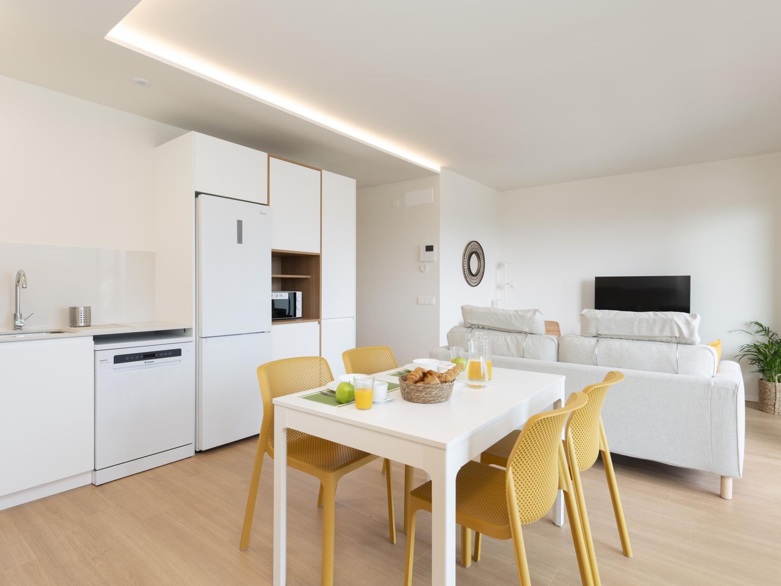 Una cucina moderna con mobili bianchi, lavastoviglie integrata e zona pranzo con tavolo bianco e sedie gialle.