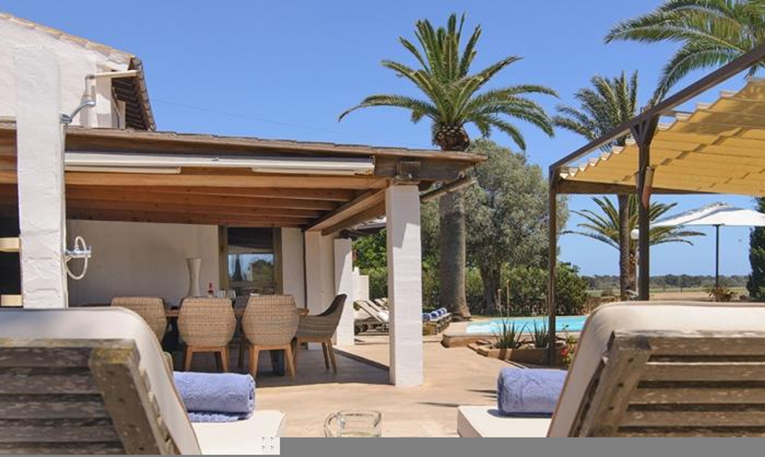 Ein überdachter Außenbereich mit Korbstühlen, einem Tisch, einem Pool und Palmen im Hintergrund.