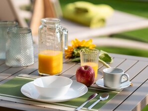 En frukostuppsättning på ett utomhusbord med en kanna apelsinjuice, ett glas apelsinjuice, en skål, en kopp och en tallrik.