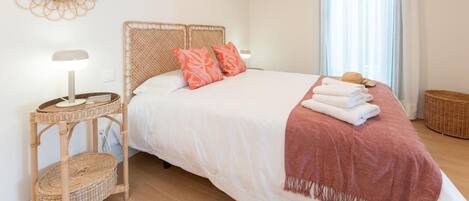 A neatly arranged bedroom with a bed, a bedside table with a lamp, a wicker side table, and a window with curtains.
