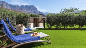 A garden with two lounge chairs, one with a blue towel and a straw hat, and a small wooden structure in the background.