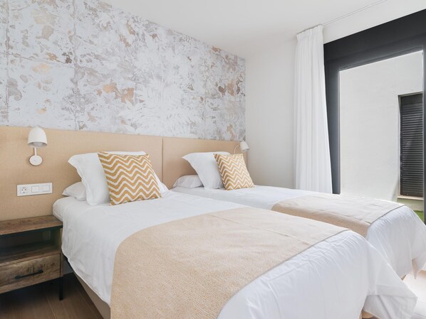 Habitación de hotel con dos camas, una mesita de noche de madera, una lámpara de pared y un amplio ventanal con cortinas.