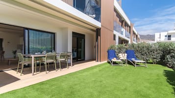 A modern outdoor patio with a dining table and chairs, a grassy lawn, and two sun loungers.