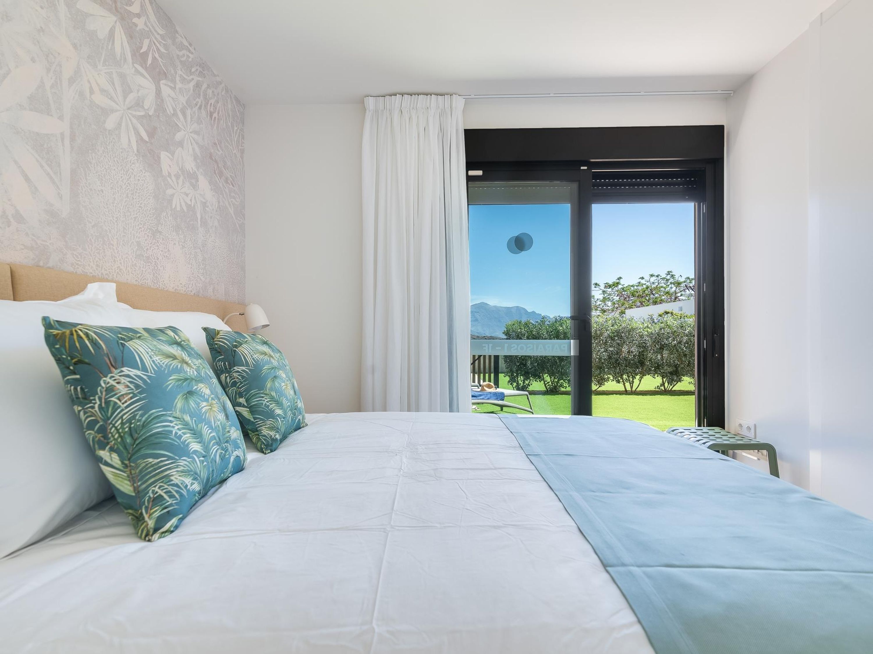 A bedroom with a bed, two pillows, a nightstand, a window with a view of a garden and mountains, and a sliding glass door.