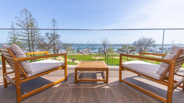 Un balcon avec deux chaises en bois et une petite table, donnant sur une marina où sont amarrés des bateaux, et sous un ciel dégagé.