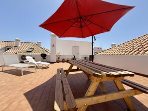 A patio with a wooden table and benches, a red umbrella, and a grill. - Casa Mergoux Maravilhoso Apartamento em Tavira (Tavira)