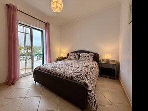 A bedroom with a bed, two bedside tables with lamps, a large window with curtains, and a view of the outside. - Casa Mergoux Maravilhoso Apartamento em Tavira (Tavira)