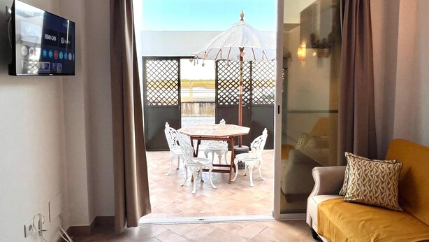 A room with a TV, a sofa, a dining table with chairs, and a view through a glass door to an outdoor area with a white gazebo. - Refugio da Ria 3 Mins da Praia p tio Central (Tavira)