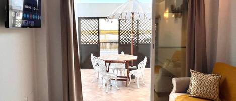 A room with a TV, a sofa, a dining table with chairs, and a view through a glass door to an outdoor area with a white gazebo.