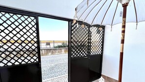A room with a wooden table, a white umbrella, and a view of a cobblestone street through a black lattice door. - Refugio da Ria 3 Mins da Praia p tio Central (Tavira)