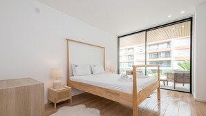 A modern bedroom with a wooden four-poster bed, a white rug, a nightstand with a lamp, and a view of a building through large windows. - Waterfront Luxury On Ria Formosa (Olhao)