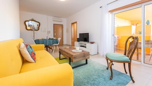A living room with a yellow sofa, a wooden coffee table, a TV, and a dining area with a table and chairs. - Casa Nobre Magnifico Apartamento com Piscina (Tavira)