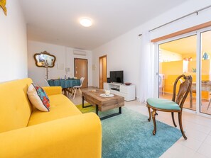 Ein Wohnzimmer mit einem gelben Sofa, einem Holztisch, einem Fernseher und einem Essbereich mit Tisch und Stühlen.