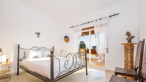 A bedroom with a metal bed frame, white bedding, a small bedside table with a lamp, a wall clock, and a view through a glass door to an outdoor seating area. - Casa Nobre Magnifico Apartamento com Piscina (Tavira)