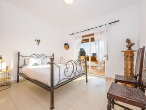 Ein Schlafzimmer mit Metallbettgestell, weißer Bettwäsche, einem kleinen Nachttisch mit Lampe, einer Wanduhr und Blick durch eine Glastür auf einen Außenbereich mit Sitzgelegenheiten.