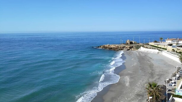 A coastal beach scene with clear skies, a rocky outcrop, and a row of buildings along the shore.