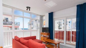 A spacious living room with a red sofa, a wooden coffee table, and a TV on a stand. Large windows offer a view of the sea and buildings.