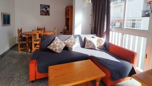 A furnished room with a sofa, a wooden table, a dining table with chairs, a cabinet, and a window with curtains.