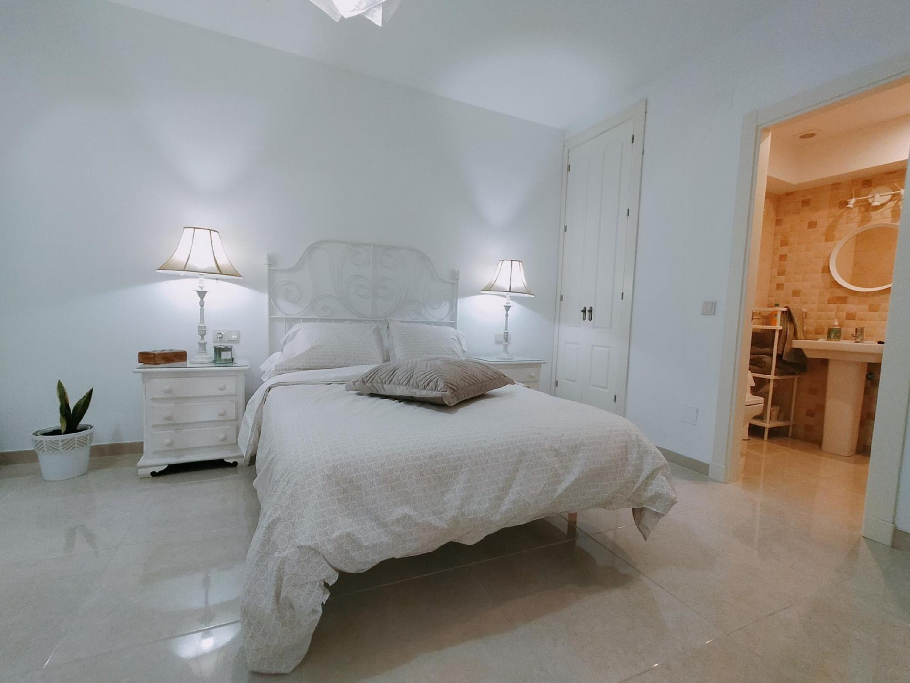 Une chambre à coucher avec un lit, deux tables de chevet, une commode, une plante en pot et une salle de bain visible à travers une porte ouverte.